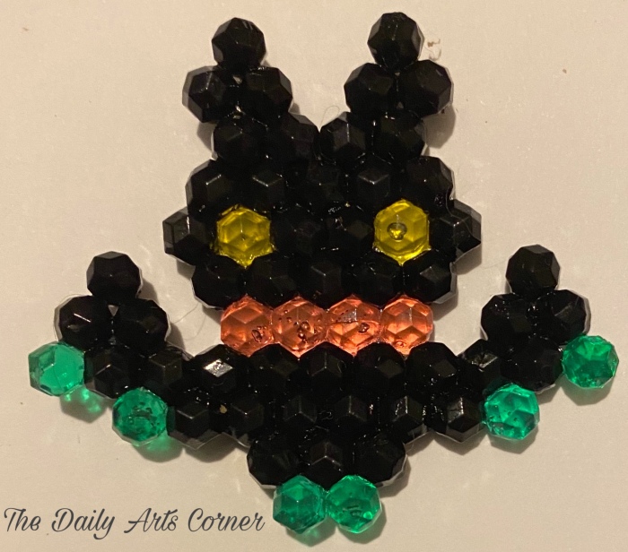 Black Aquabeads Polygon Bat with yellow eyes, green feet and hands, a pink line around the mouth.